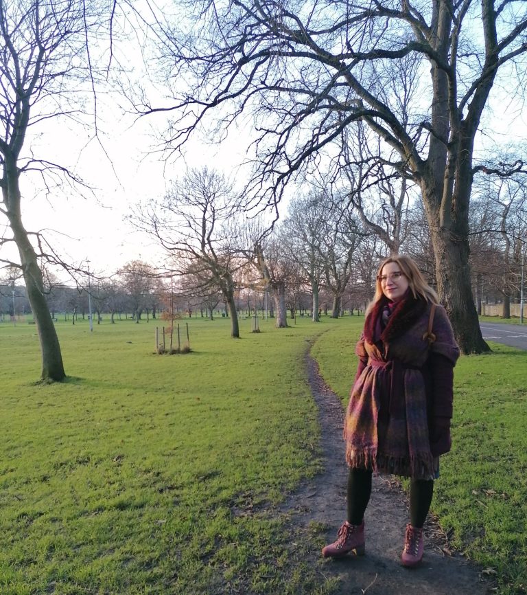 Zsofia Kaplar therapist and coach Image of a smiling Zsofia Kaplar, sitting on a wicker chair with a notebook next to plants